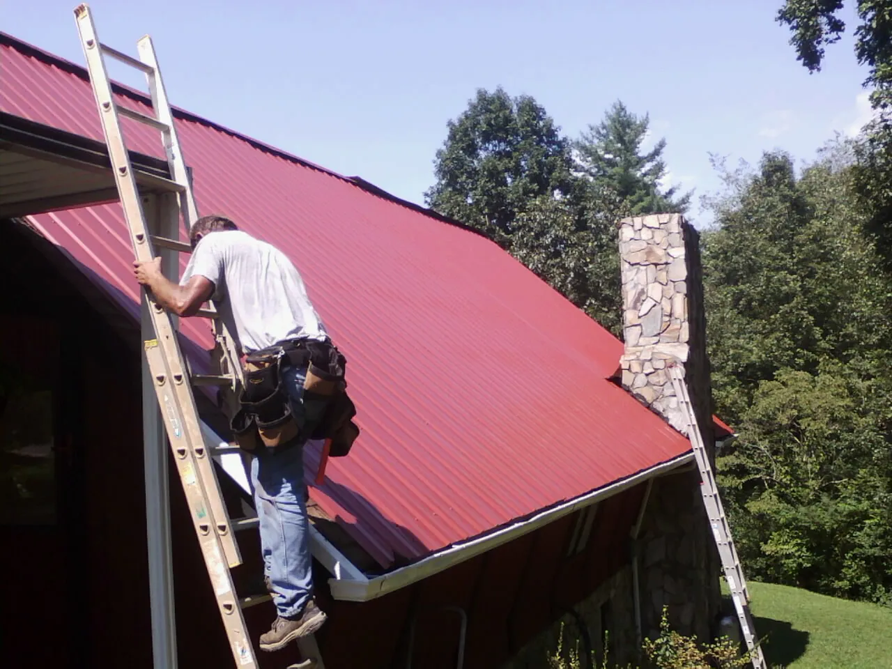 Roofer on a ladder beside a red metal roof and stone chimney — Soffit & Fascia Repair by Roofing Brothers in Western Springs